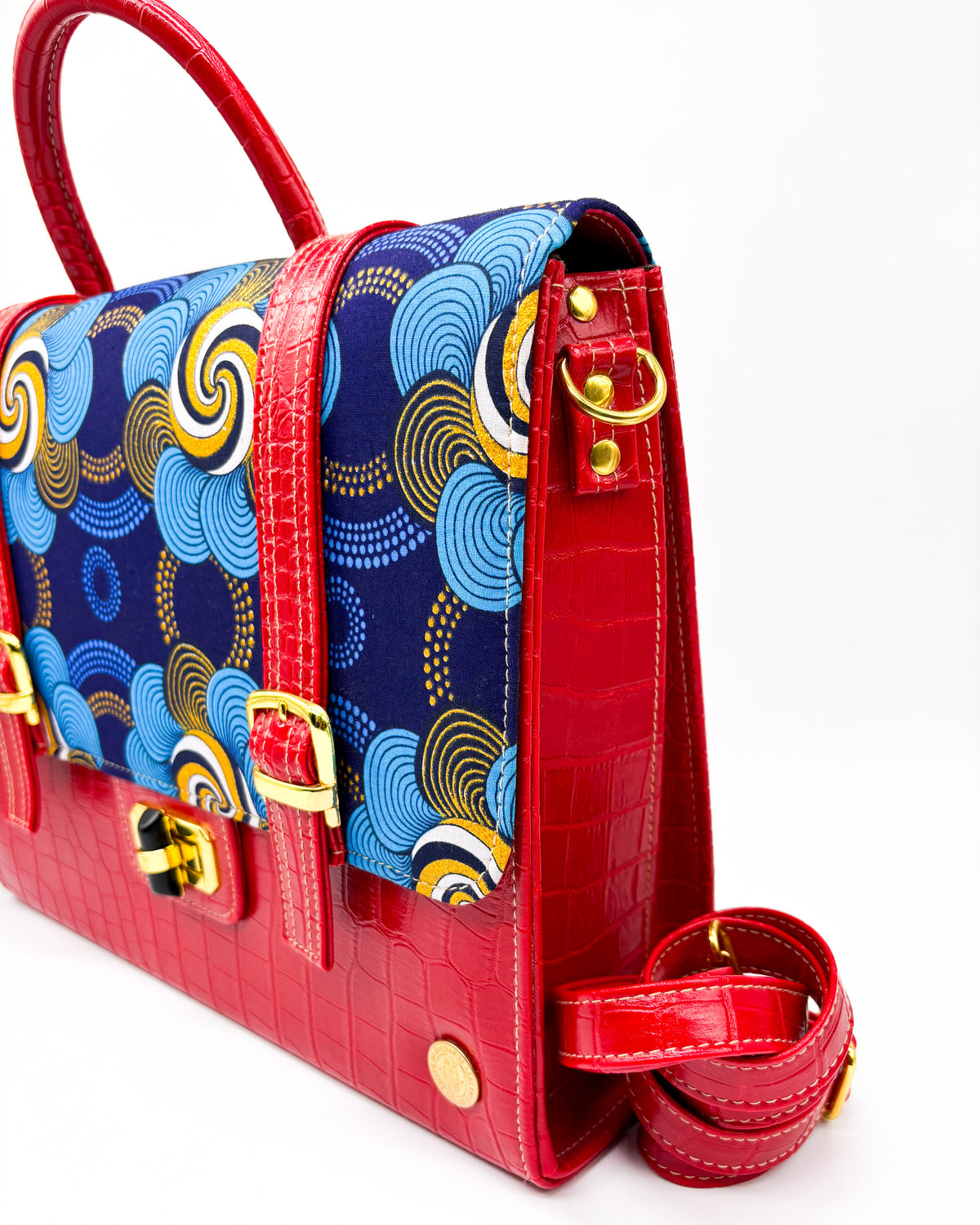 Side angle, red vegan leather handbag or backpack with gold and blue African wax fabric inlay, two gold buckled clasps and gold twist-lock.