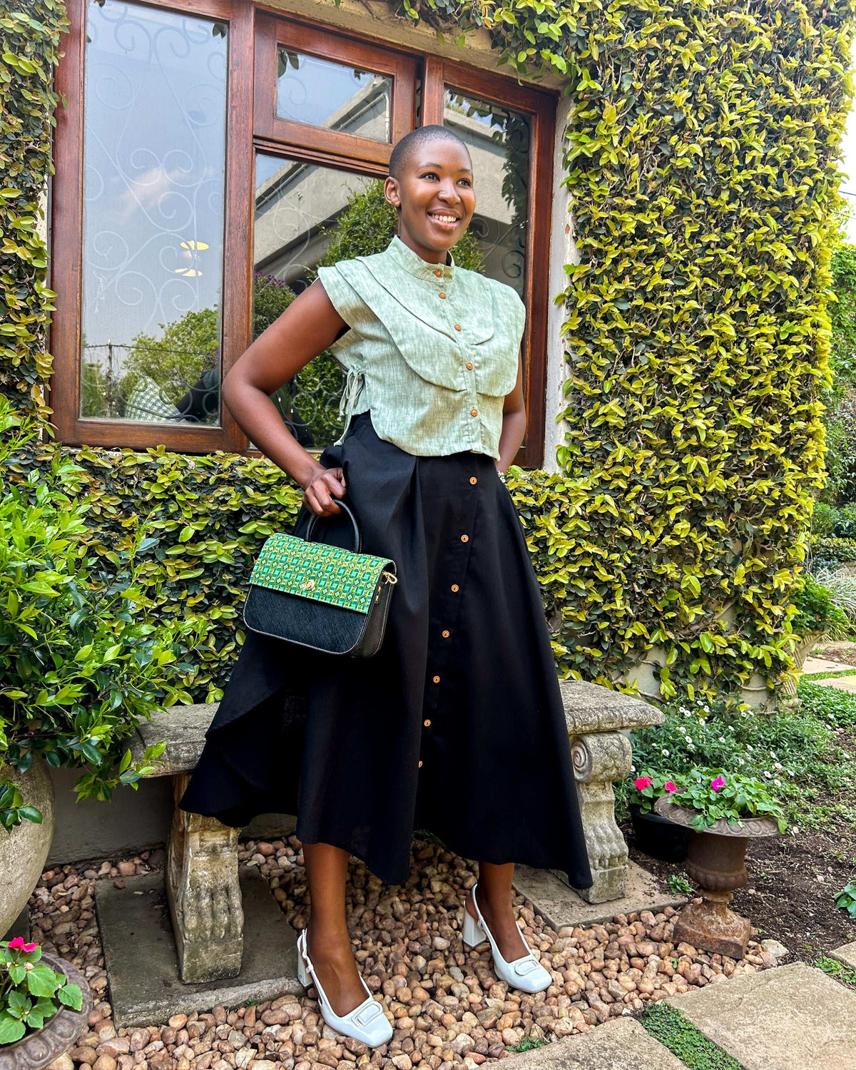 Smiling African woman wearing a Zandi Skirt in black with a Dr Pachanga handbag, and white heeled shoes.