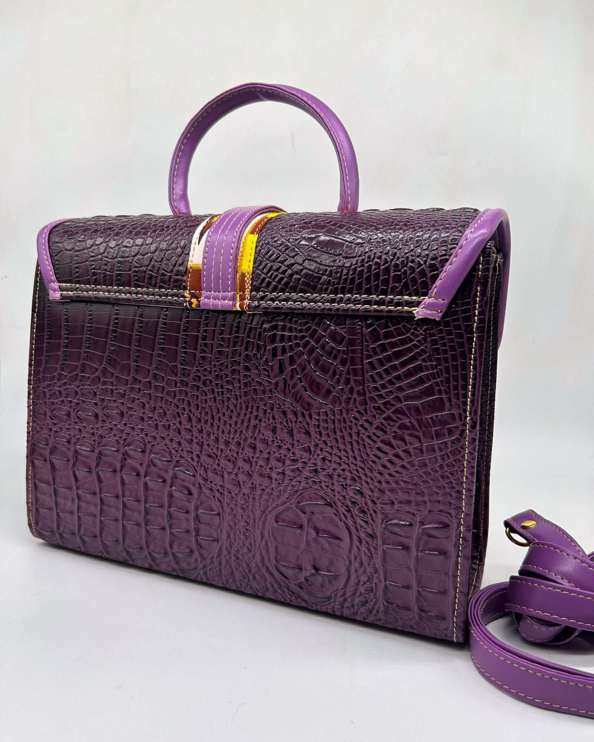 Back view, purple vegan leather handbag with yellow and brown African Wax fabric inlay, a buckled strap, gold magnetic clasp and adjustable strap. 