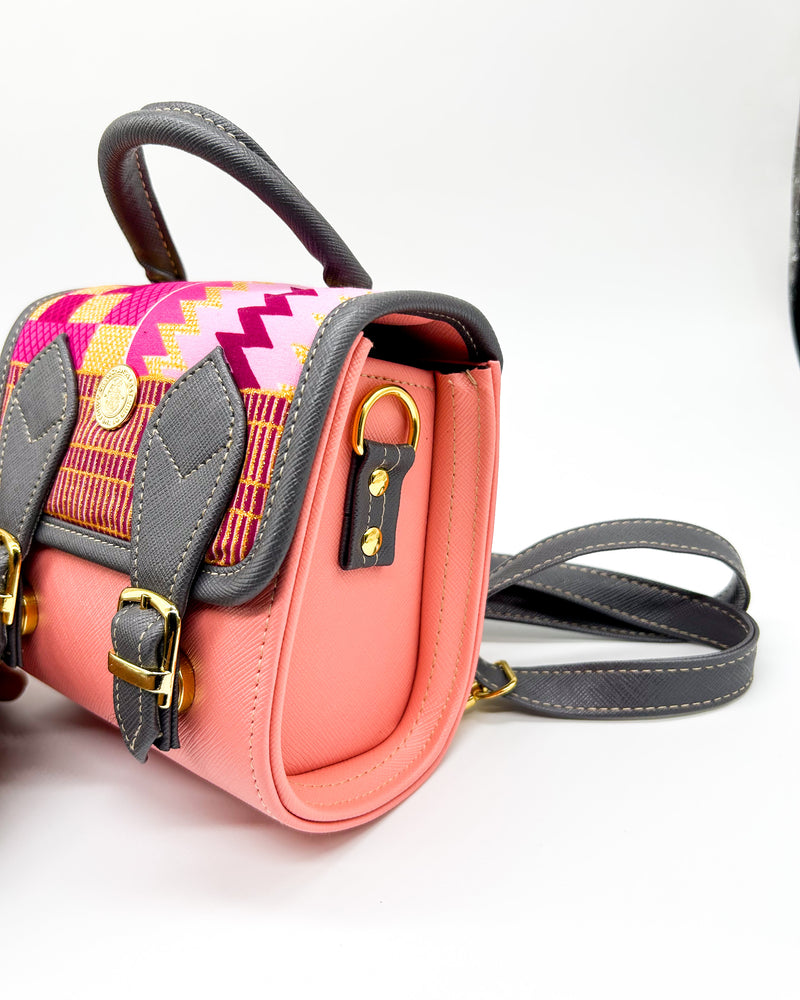 Side angle, salmon pink and black vegan leather handbag or backpack with gold African wax fabric inlay and two buckled clasps.