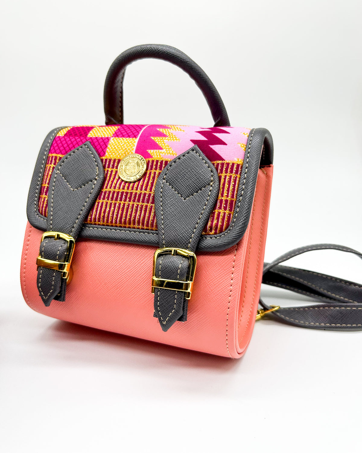 Front angle, salmon pink and black vegan leather handbag or backpack with gold African wax fabric inlay and two buckled clasps.