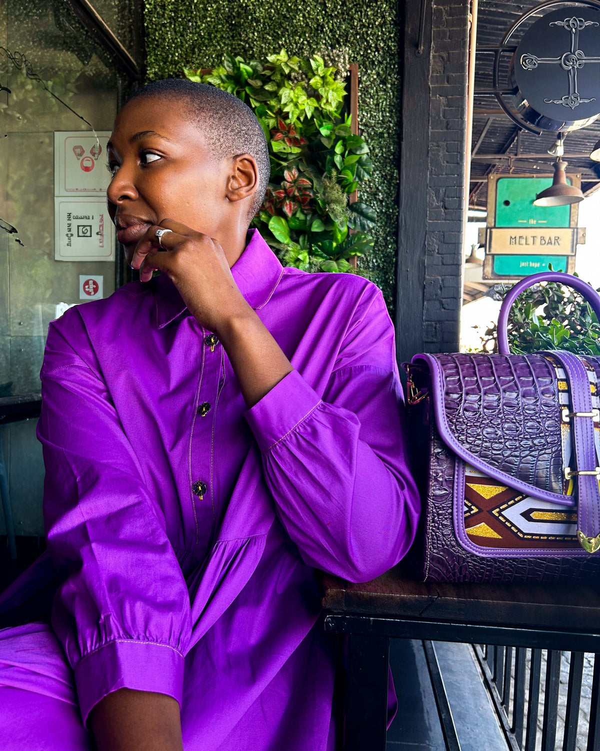 African woman wearing a Mimi Dress in purple, with a Dr Pachanga handbag on a table.