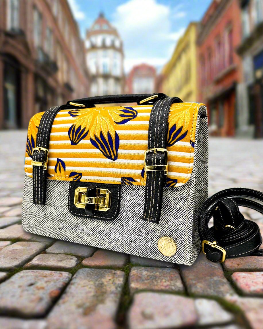 Town setting, grey vegan leather handbag with yellow and orange African Gold Wax fabric inlay, two gold buckled straps, a twist-lock clasp and adjustable strap.